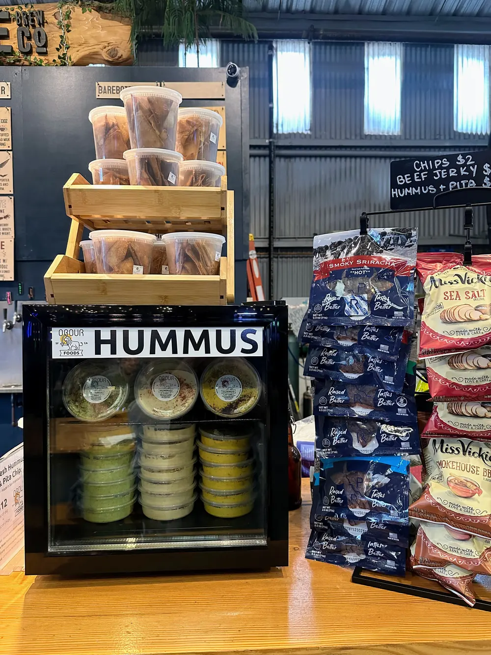 A branded Obour Hummus Fridge stocked with hummus and toum, with a crate of pita chips on top