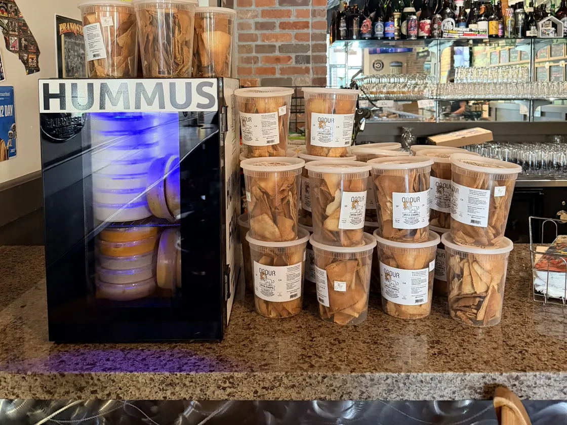 An Obour Hummus Fridge restocked, with fresh tubs of pita chips stacked beside it