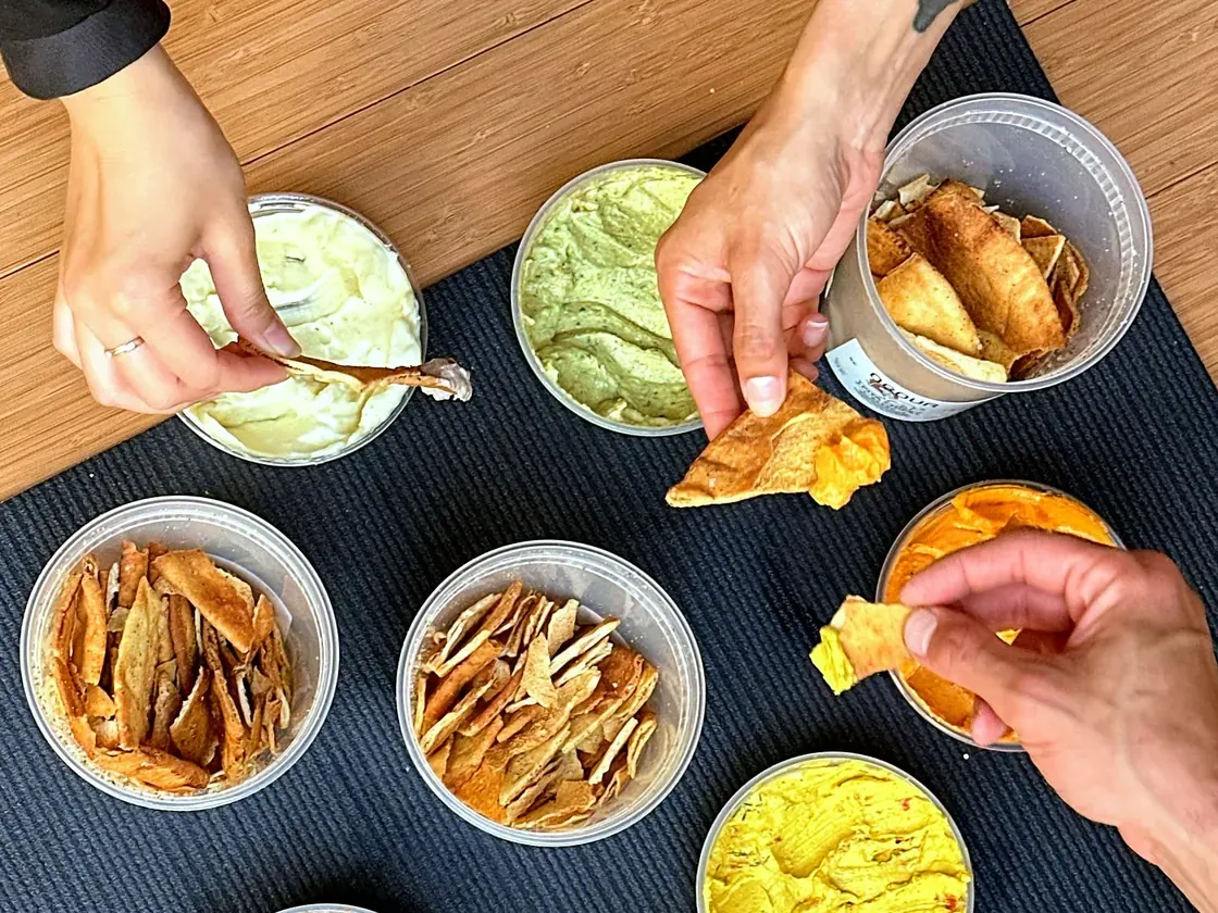 Hands dipping pita chips into round tubs of Obour hummus and toum